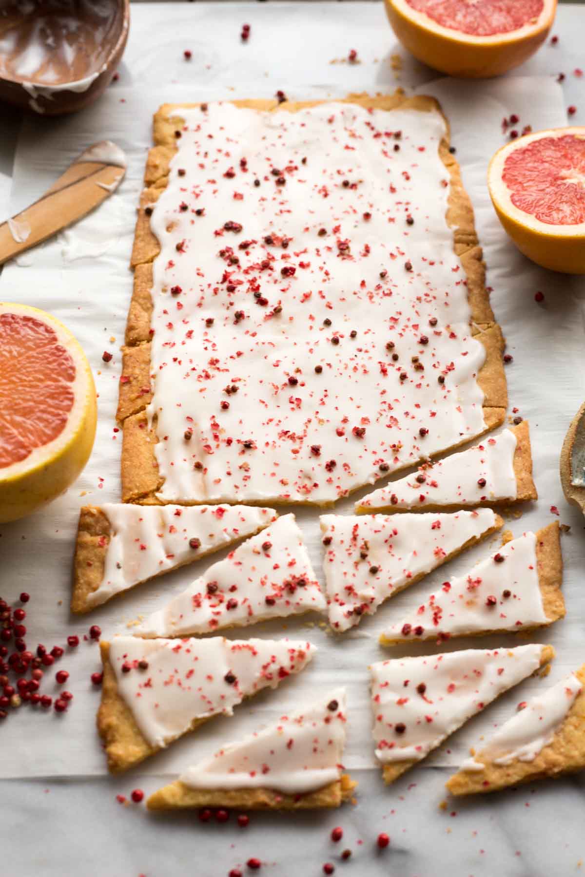 A sheet of glazed grapefruit shortbread cut into long triangles, sprinkled with pink peppercorns and arranged on parchment paper with grapefruit halves nearby.