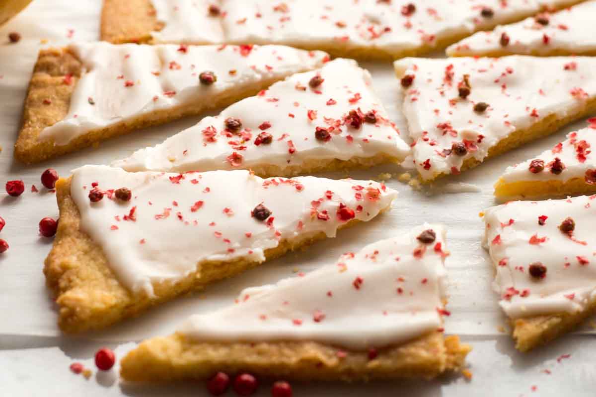 Iced grapefruit shortbread triangles scattered across parchment paper, each piece topped with crushed pink peppercorns for texture and color.