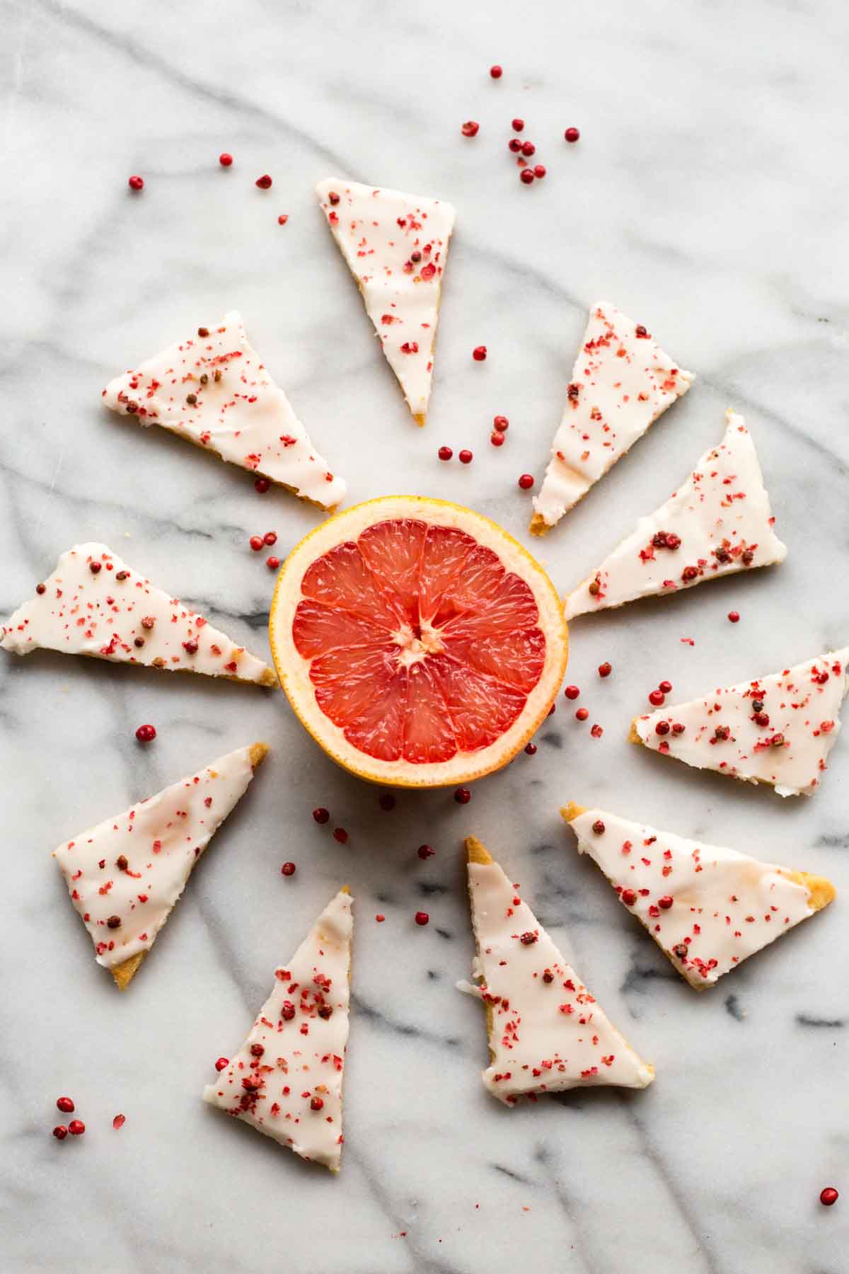 A halved ruby red grapefruit in the center with iced grapefruit shortbread triangles arranged in a circular pattern around it, sprinkled with pink peppercorns.