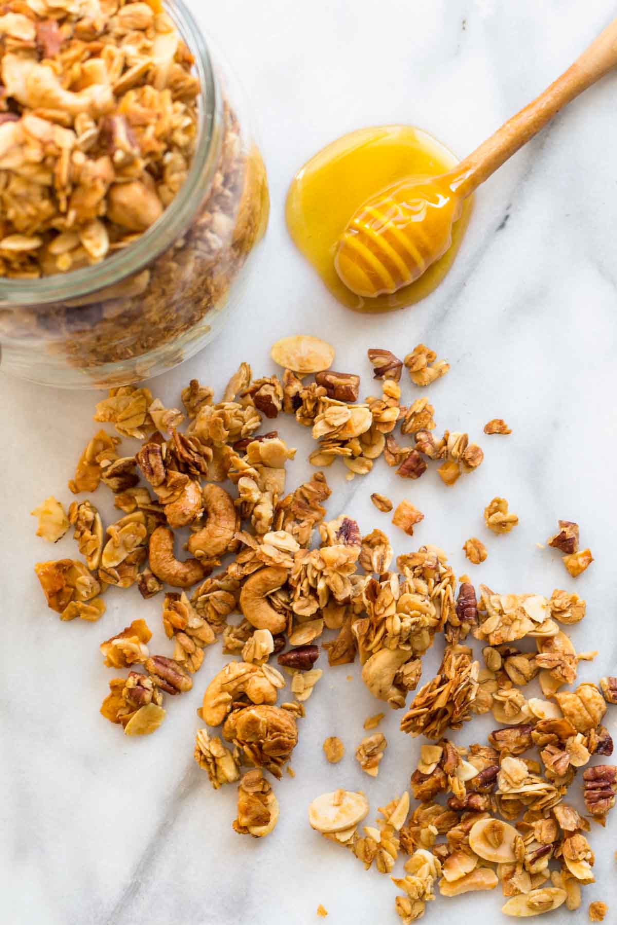Crunchy homemade granola scattered on a marble surface next to a glass jar and a honey dipper resting in a small pool of honey.