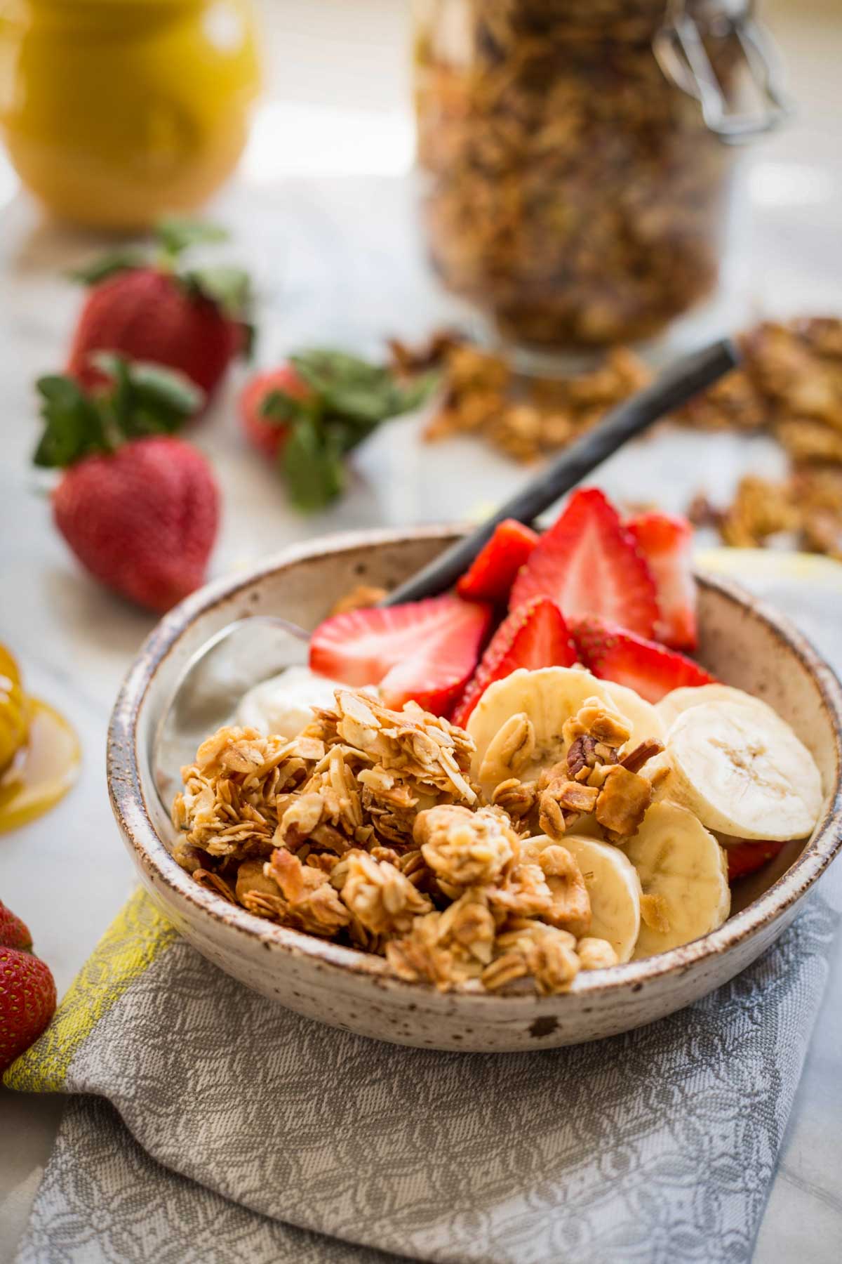 A ceramic bowl filled with yogurt, sliced bananas, strawberries and a generous scoop of homemade granola, with extra berries and a jar of granola in the background.