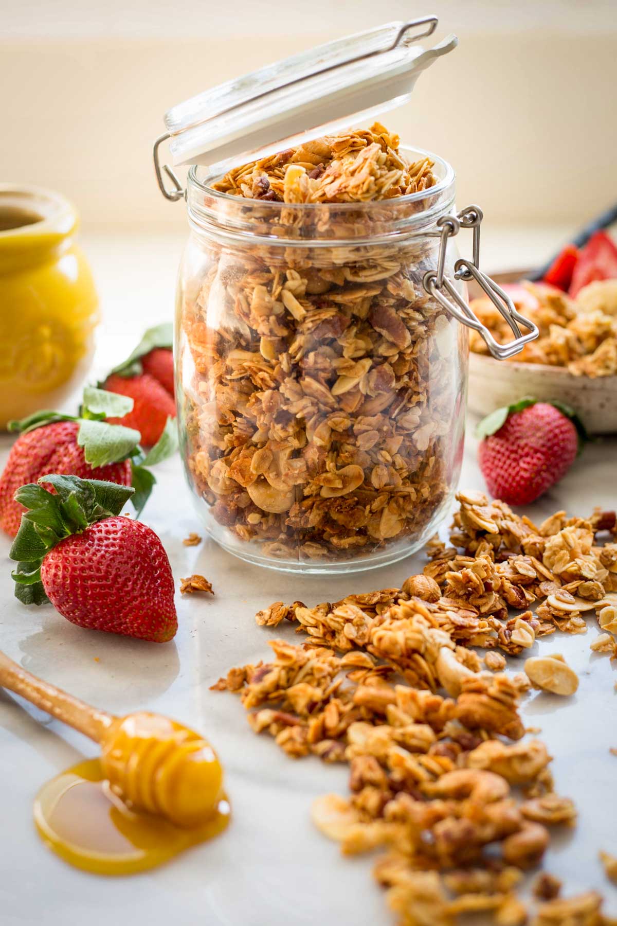 A clear latch-top jar overflowing with golden homemade granola surrounded by fresh strawberries and scattered clusters on a marble countertop.