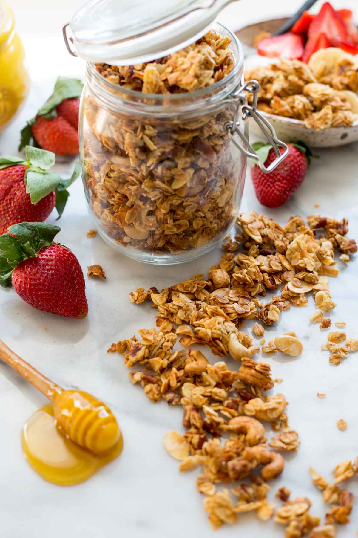 An open jar of homemade granola on a marble surface with fresh strawberries, loose granola pieces and a honey dipper coated in honey nearby.