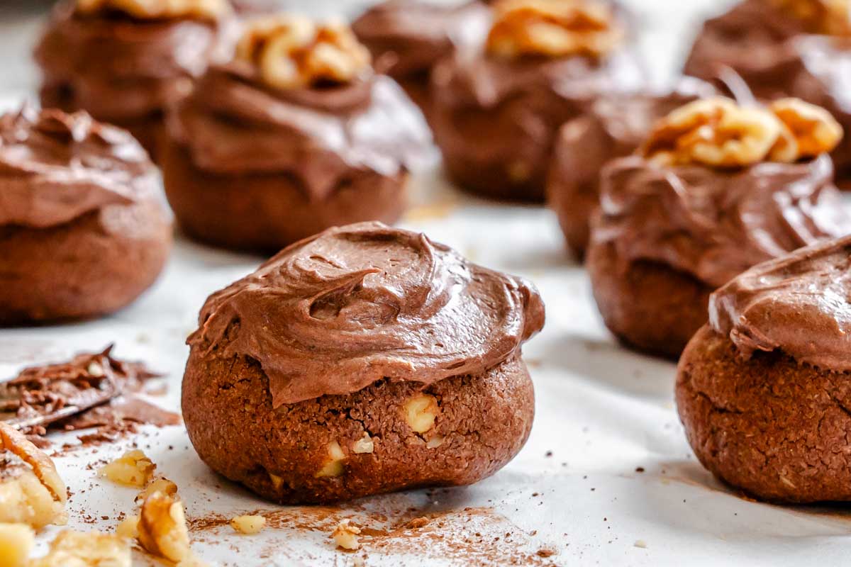 A close up of a chocolate spice cookie topped with thick chocolate frosting and chopped walnuts on parchment paper.