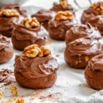 A frosted chocolate spice cookie topped with a walnut half, surrounded by more cookies in soft focus.