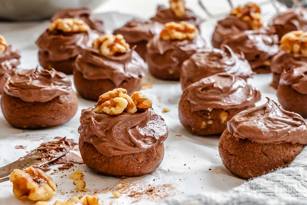 A frosted chocolate spice cookie topped with a walnut half, surrounded by more cookies in soft focus.