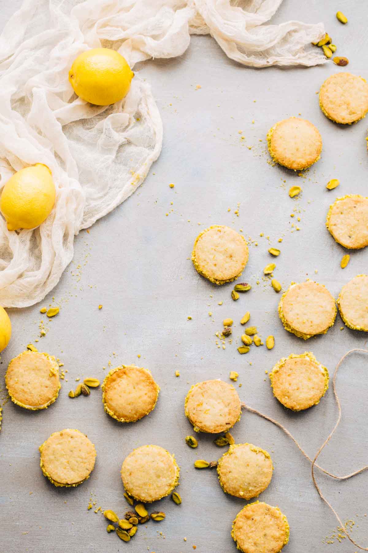 Overhead view of lemon pistachio sandwich cookies arranged on a light surface with scattered pistachios and fresh lemons.