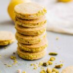 Stack of lemon pistachio sandwich cookies with chopped pistachio edges and blurred lemons in the background.