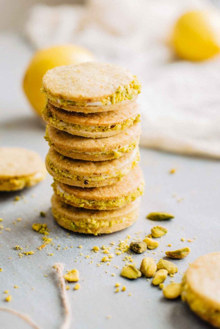 Stack of lemon pistachio sandwich cookies with chopped pistachio edges and blurred lemons in the background.