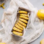 Box filled with lemon pistachio sandwich cookies nestled in parchment, surrounded by lemons and rustic fabric.