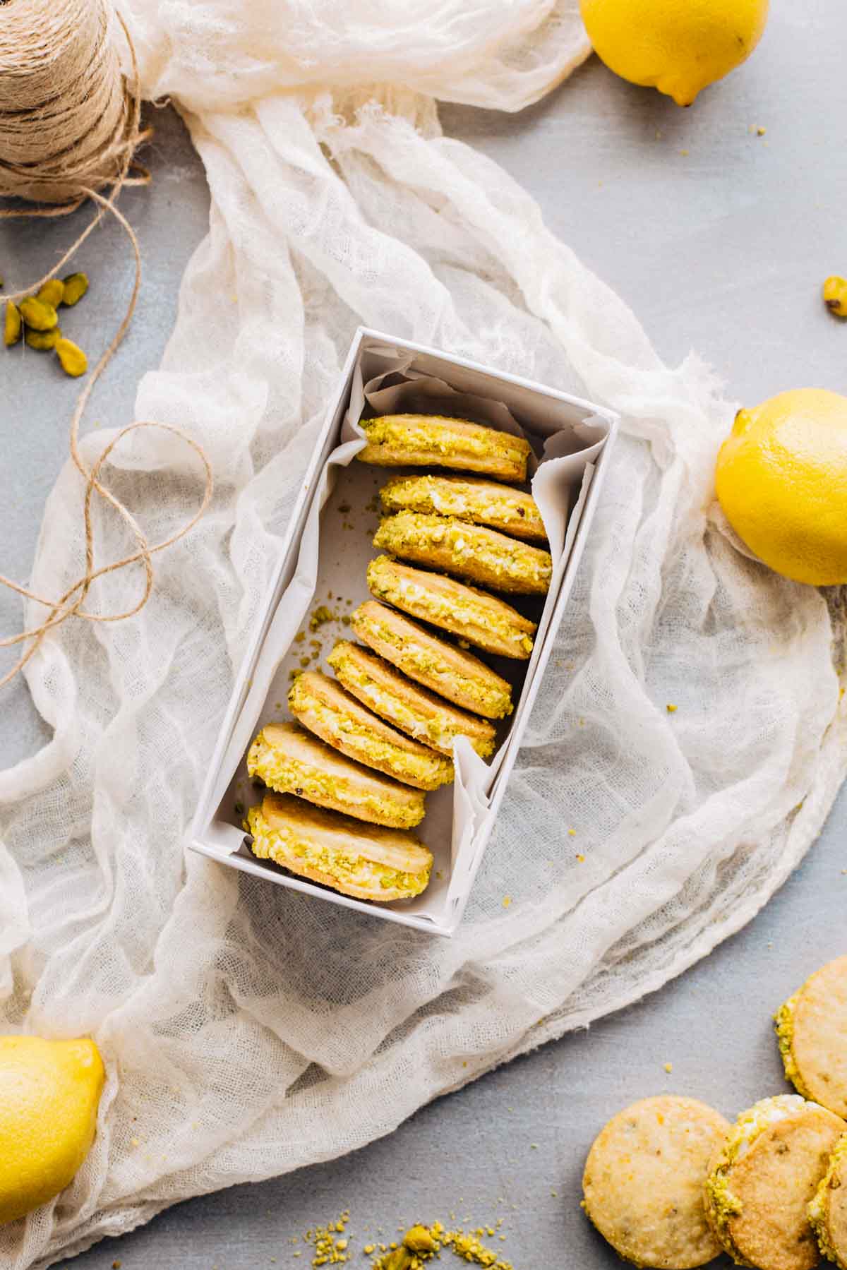 Box filled with lemon pistachio sandwich cookies nestled in parchment, surrounded by lemons and rustic fabric.