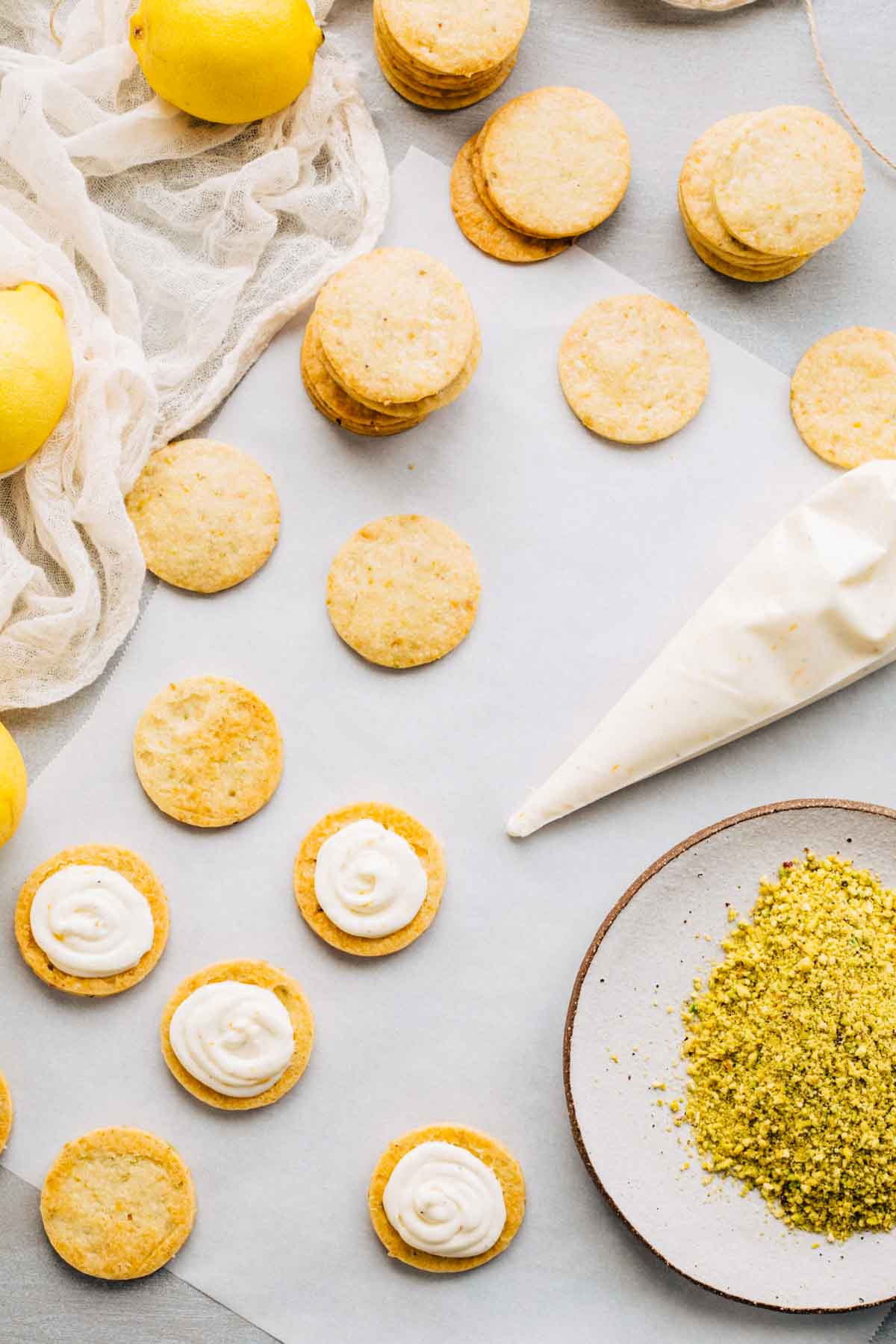 Process shot showing cookies being filled with lemon cream and rolled in crushed pistachios on a work surface.