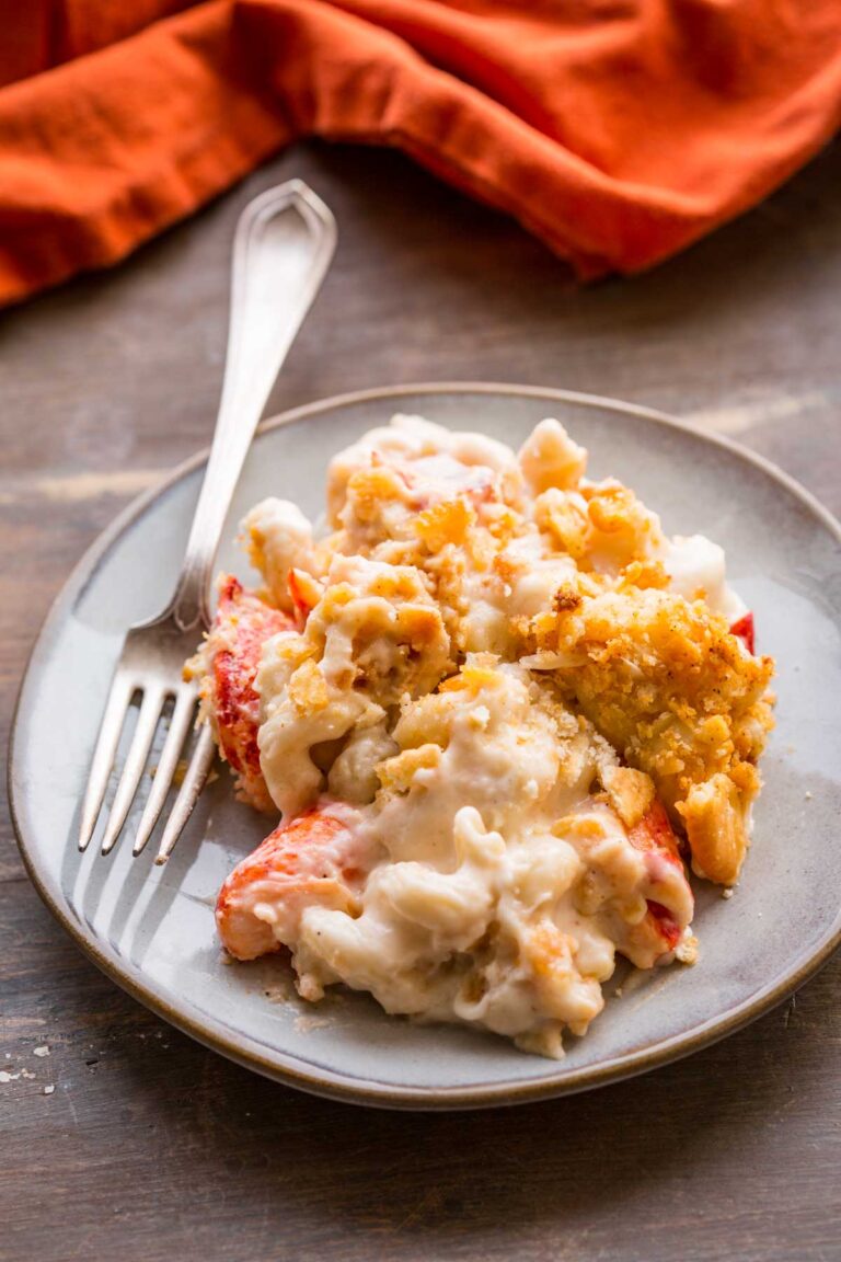 A plate of lobster mac n cheese showing large pieces of lobster, creamy pasta, and crisp cracker topping with a fork on the side.