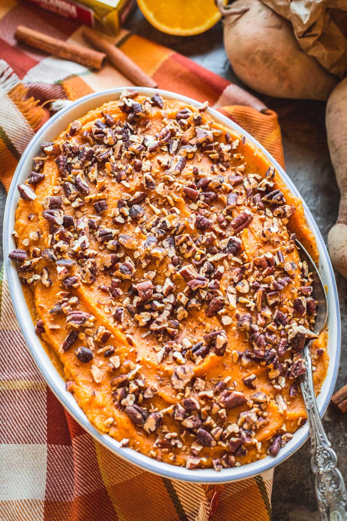 Side angle of sweet potato casserole with golden brown pecans, surrounded by oranges, cinnamon sticks, and sweet potatoes for a festive holiday setting.
