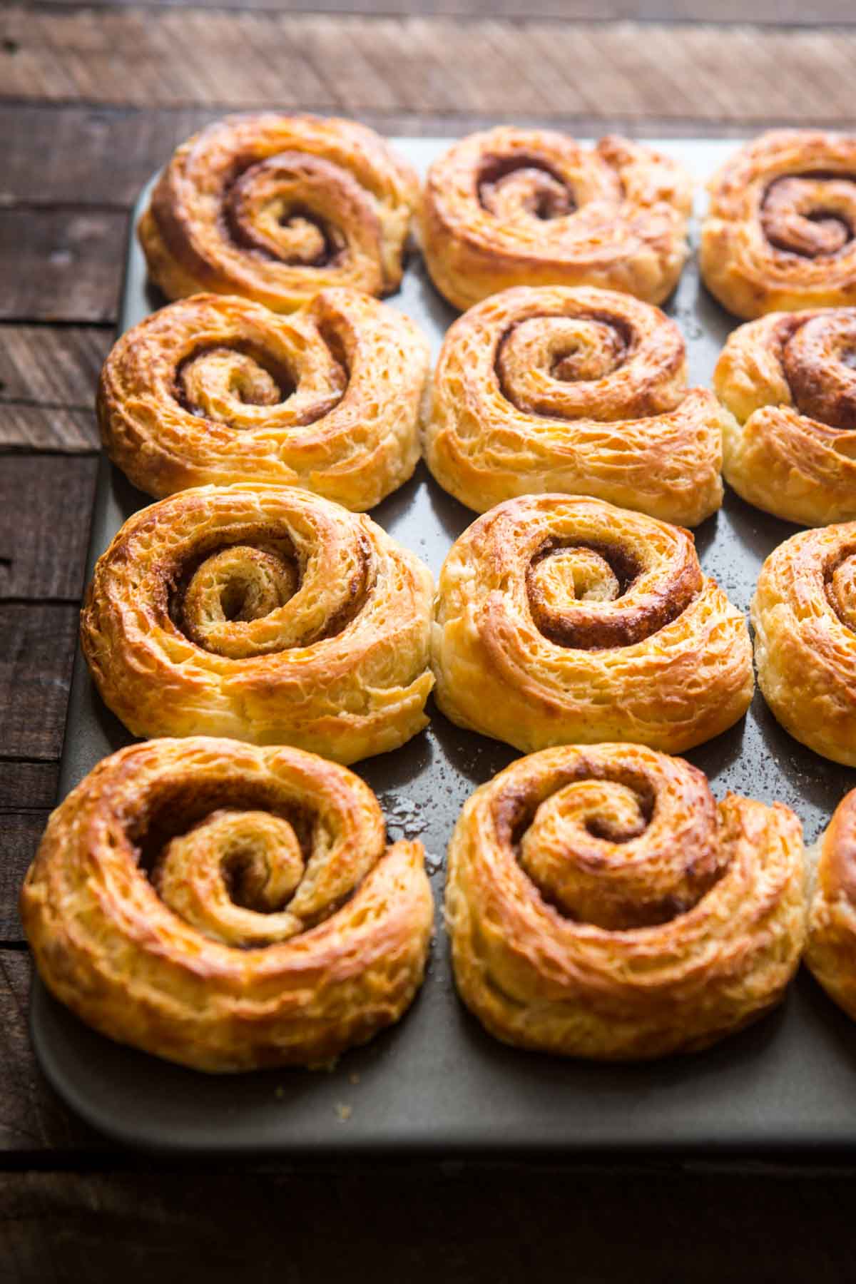 Light, flaky morning buns still in the muffin tin, showing crisp golden layers and spiraled cinnamon filling.