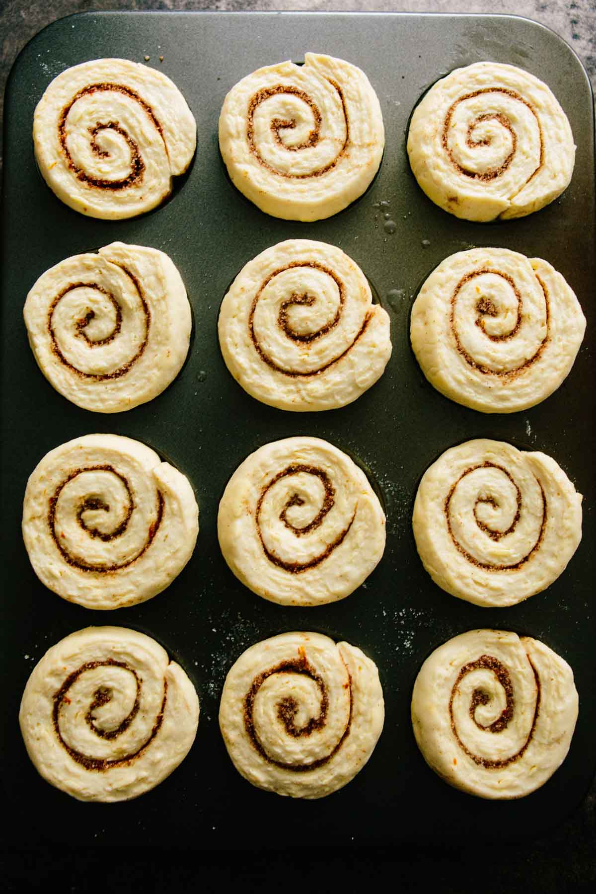 Unbaked morning buns arranged in a muffin pan, displaying their tight spirals and even cinnamon sugar swirl.