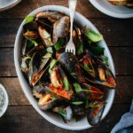 A white oval serving bowl filled with mussels marinara, garnished with fresh basil and parsley, set on a wooden table with sliced bread on the side.