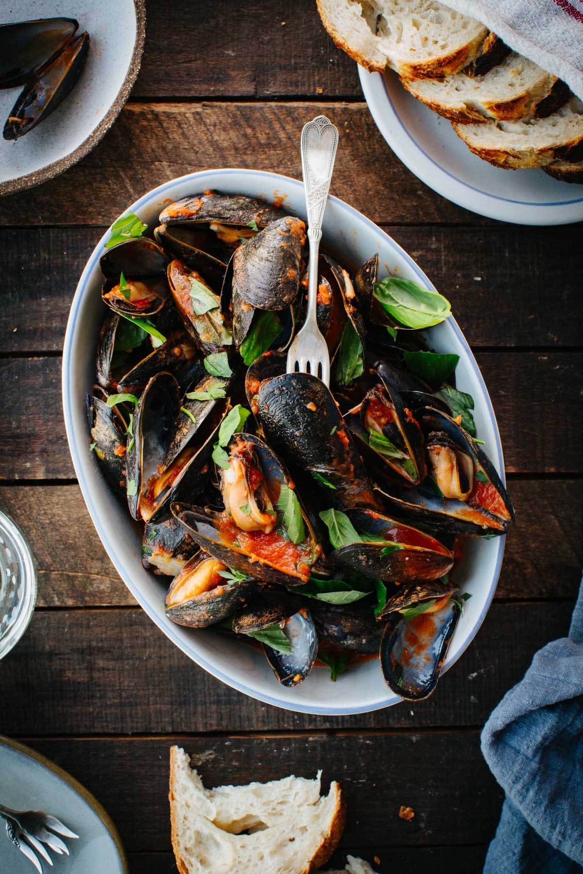 A white oval serving bowl filled with mussels marinara, garnished with fresh basil and parsley, set on a wooden table with sliced bread on the side.