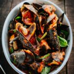 A close overhead view of a bowl piled with mussels coated in tomato sauce, topped with torn basil leaves.