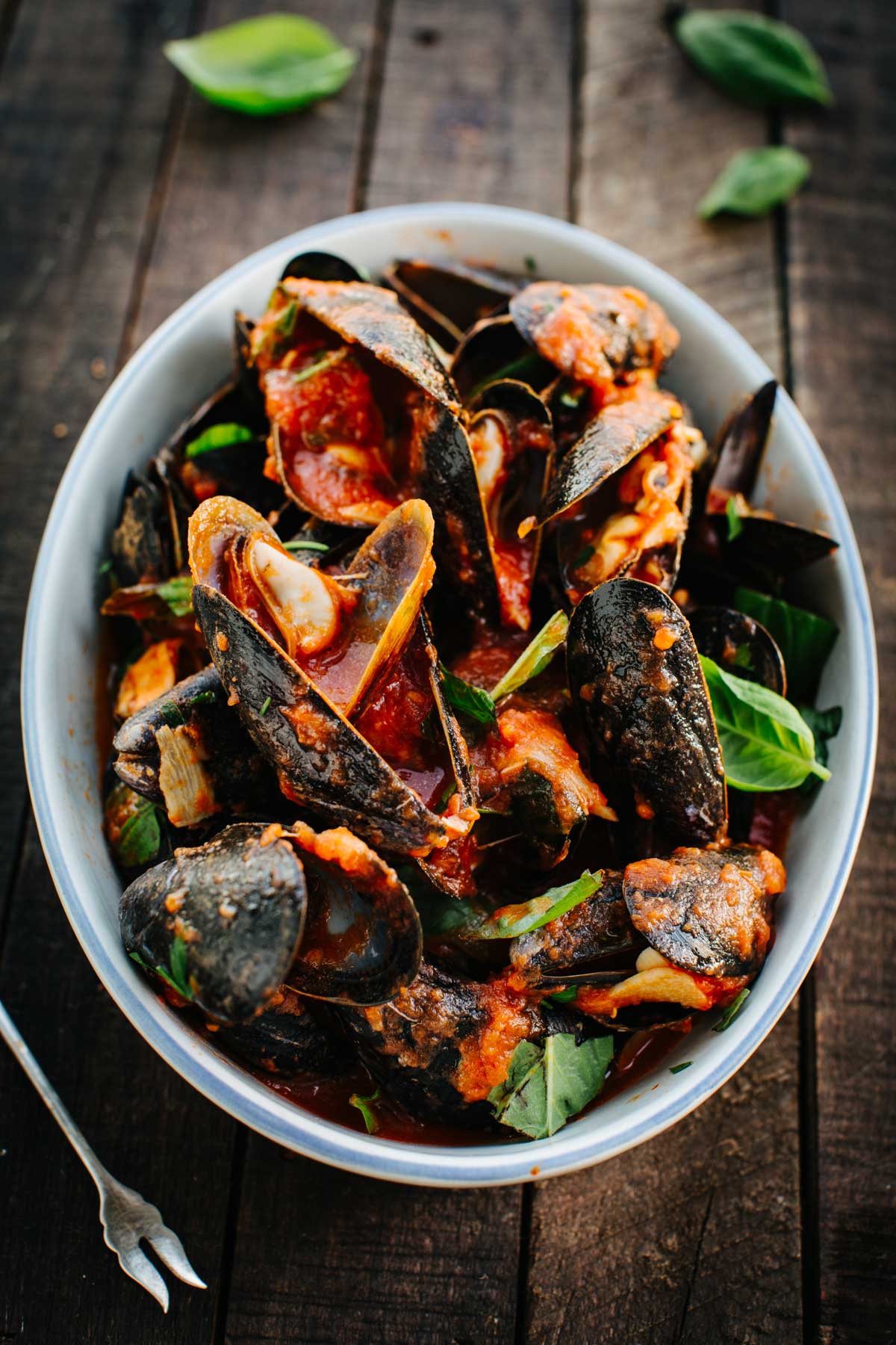 A close overhead view of a bowl piled with mussels coated in tomato sauce, topped with torn basil leaves.