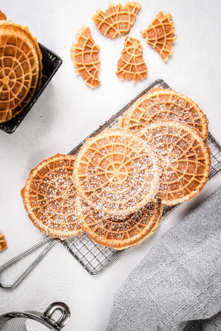 Golden pizzelles cooling on a wire rack, lightly dusted with powdered sugar, with broken cookie pieces around them.