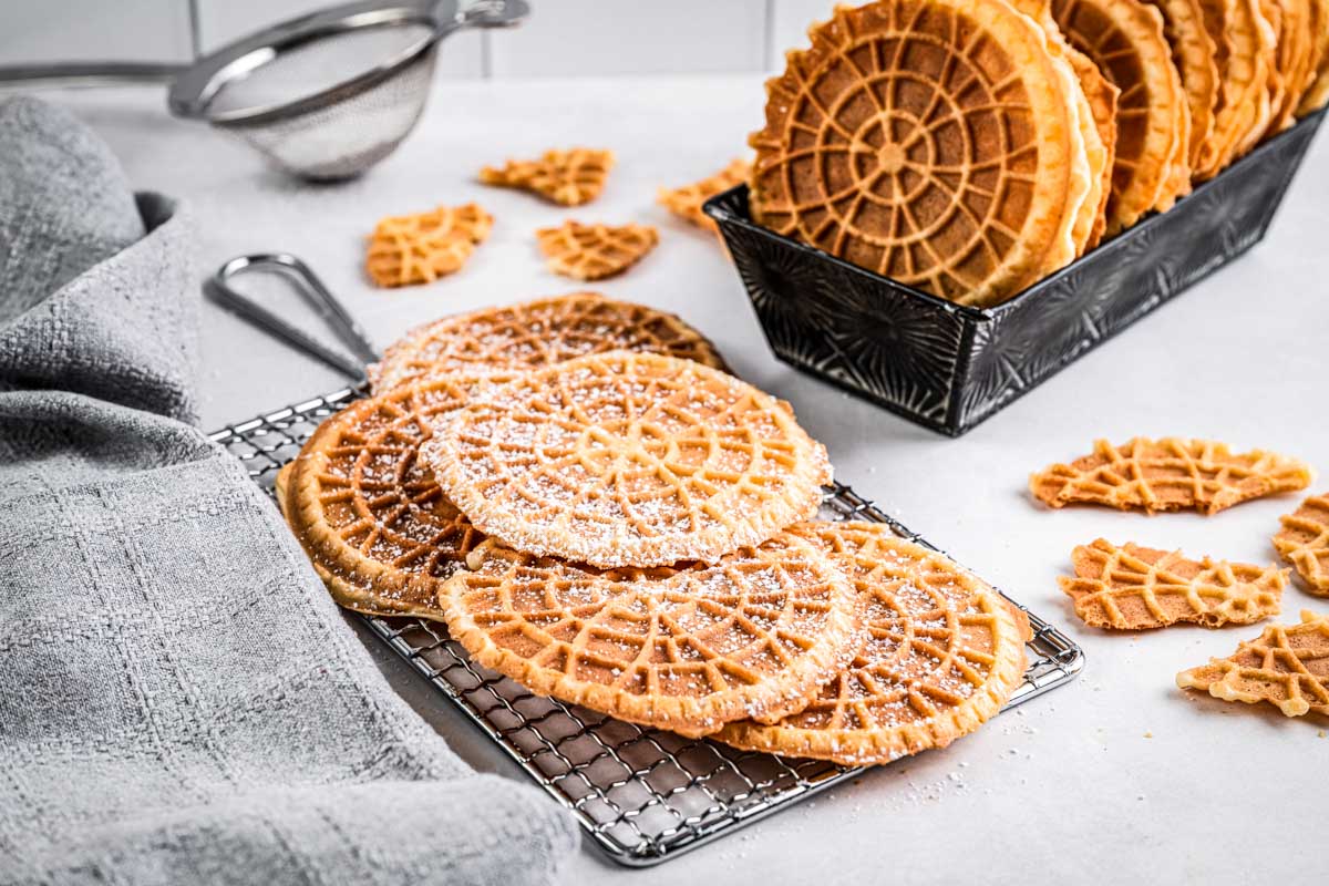 A small tray filled with stacked pizzelles next to several pizzelles cooling on a rack and scattered crisp pieces.