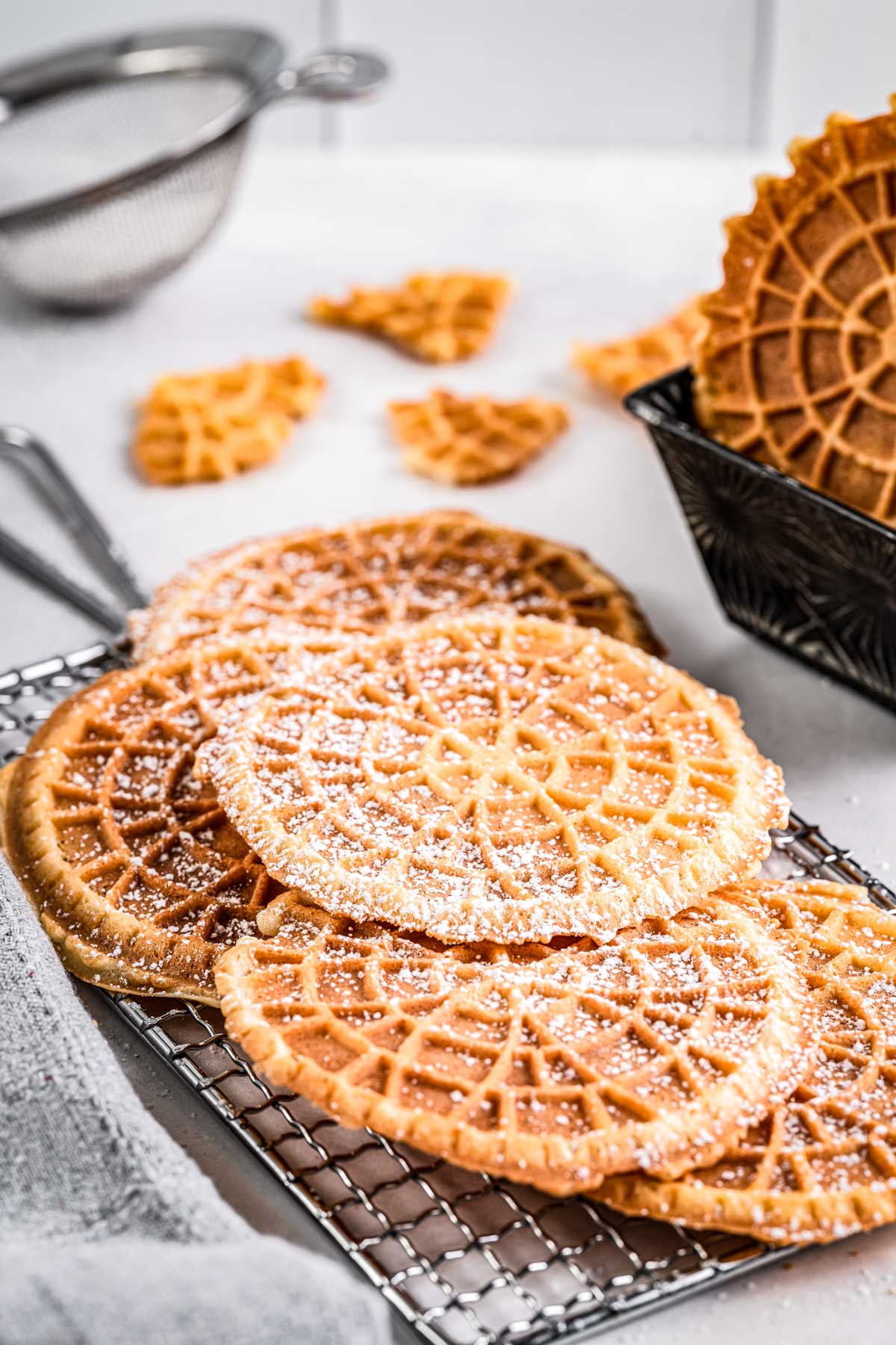 Warm pizzelles cooling on a rack with scattered broken cookie shards and a strainer in the background.