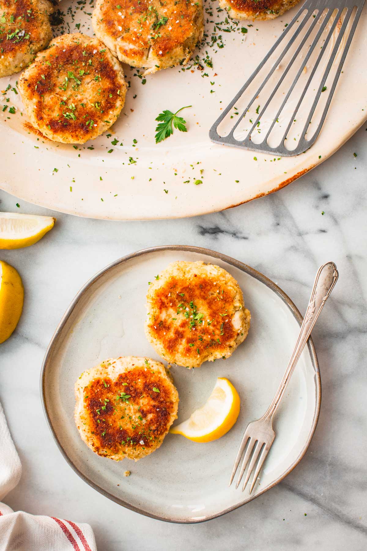 Golden brown mashed potato fish cakes on a small gray plate with lemon wedges, sprinkled with parsley, set on a marble surface with a fork.