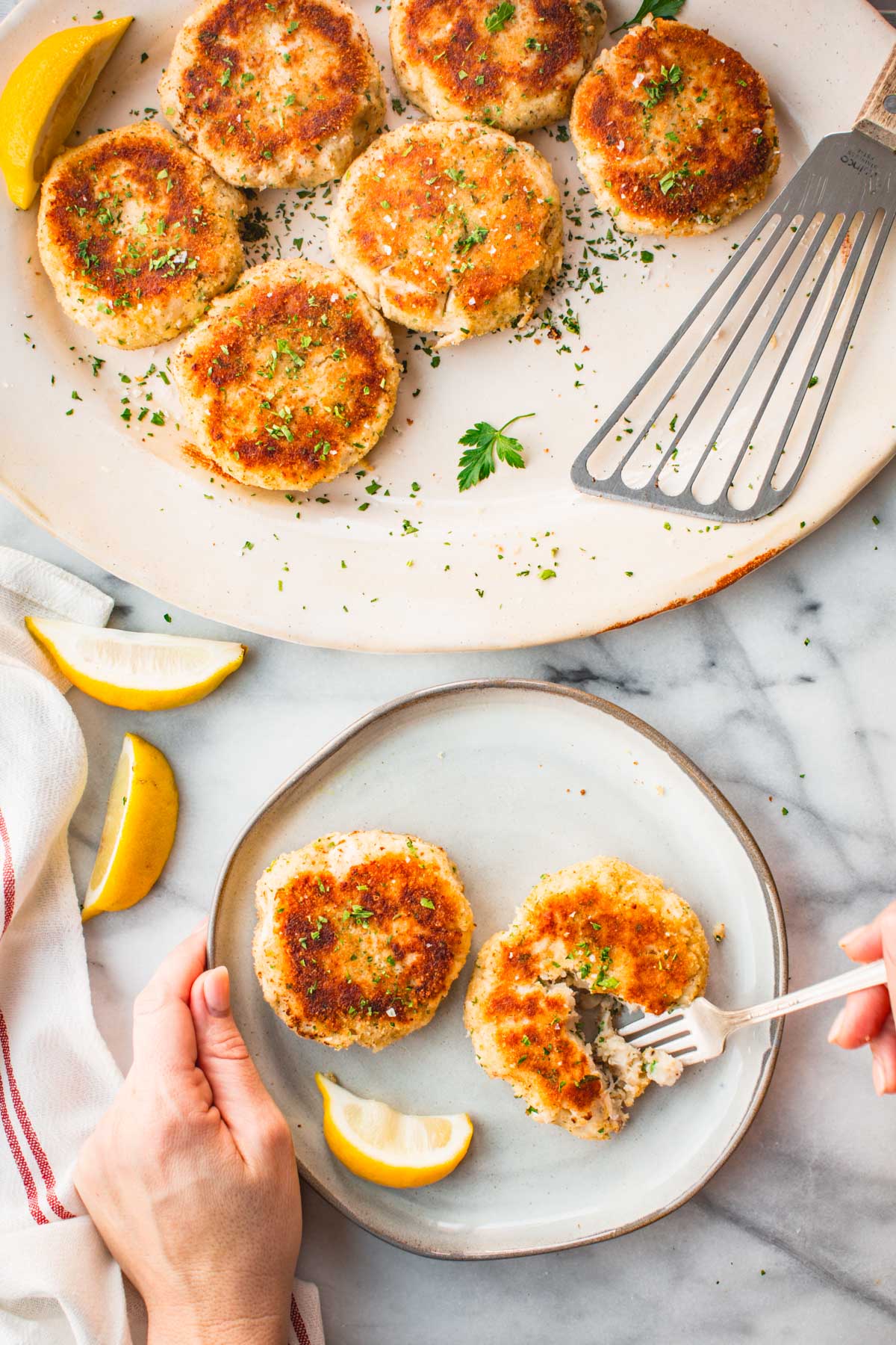 A plate of browned mashed potato fish cakes with a person cutting into one, surrounded by lemon wedges and a larger platter of fish cakes in the background.