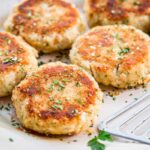Crispy mashed potato fish cakes on a large platter, topped with chopped parsley and coarse salt, with a slotted spatula nearby.