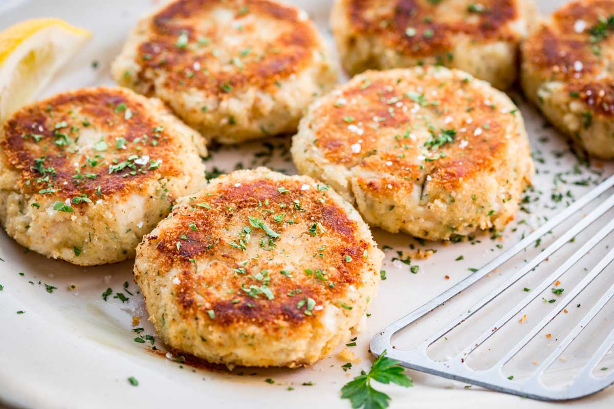 Crispy mashed potato fish cakes on a large platter, topped with chopped parsley and coarse salt, with a slotted spatula nearby.