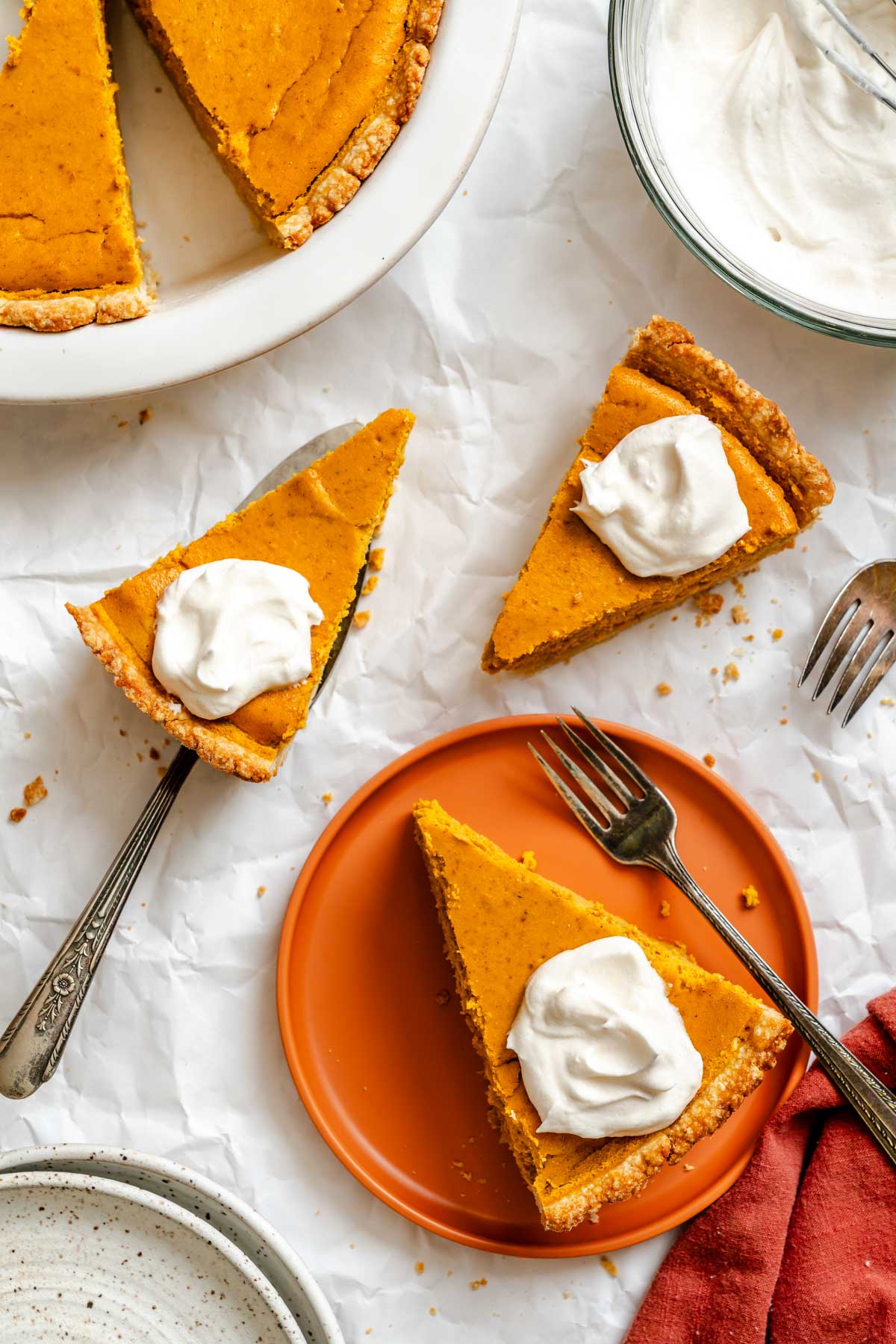 Slices of pumpkin ricotta pie served with whipped cream on an orange plate and parchment background, with forks nearby.
