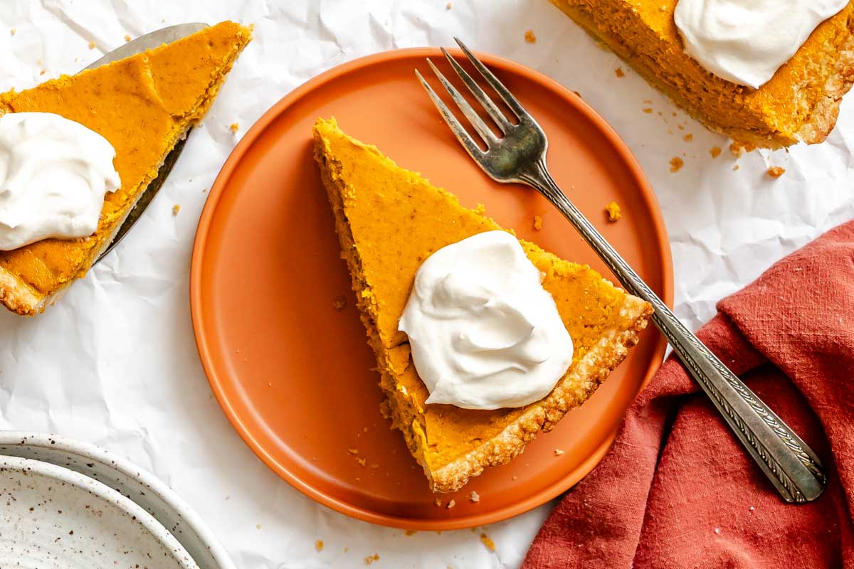 Close-up of pumpkin ricotta pie slice topped with whipped cream on an orange plate with a red napkin.