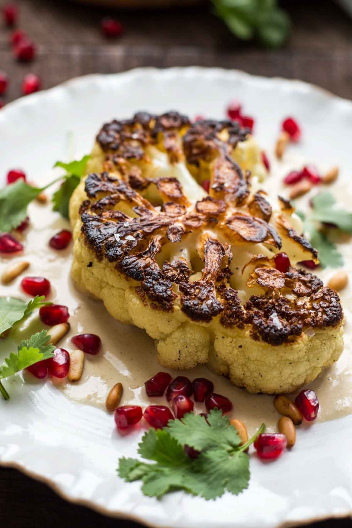 Pan-seared cauliflower steak served on a white plate over creamy tahini sauce, topped with pomegranate seeds, pine nuts, and fresh cilantro.