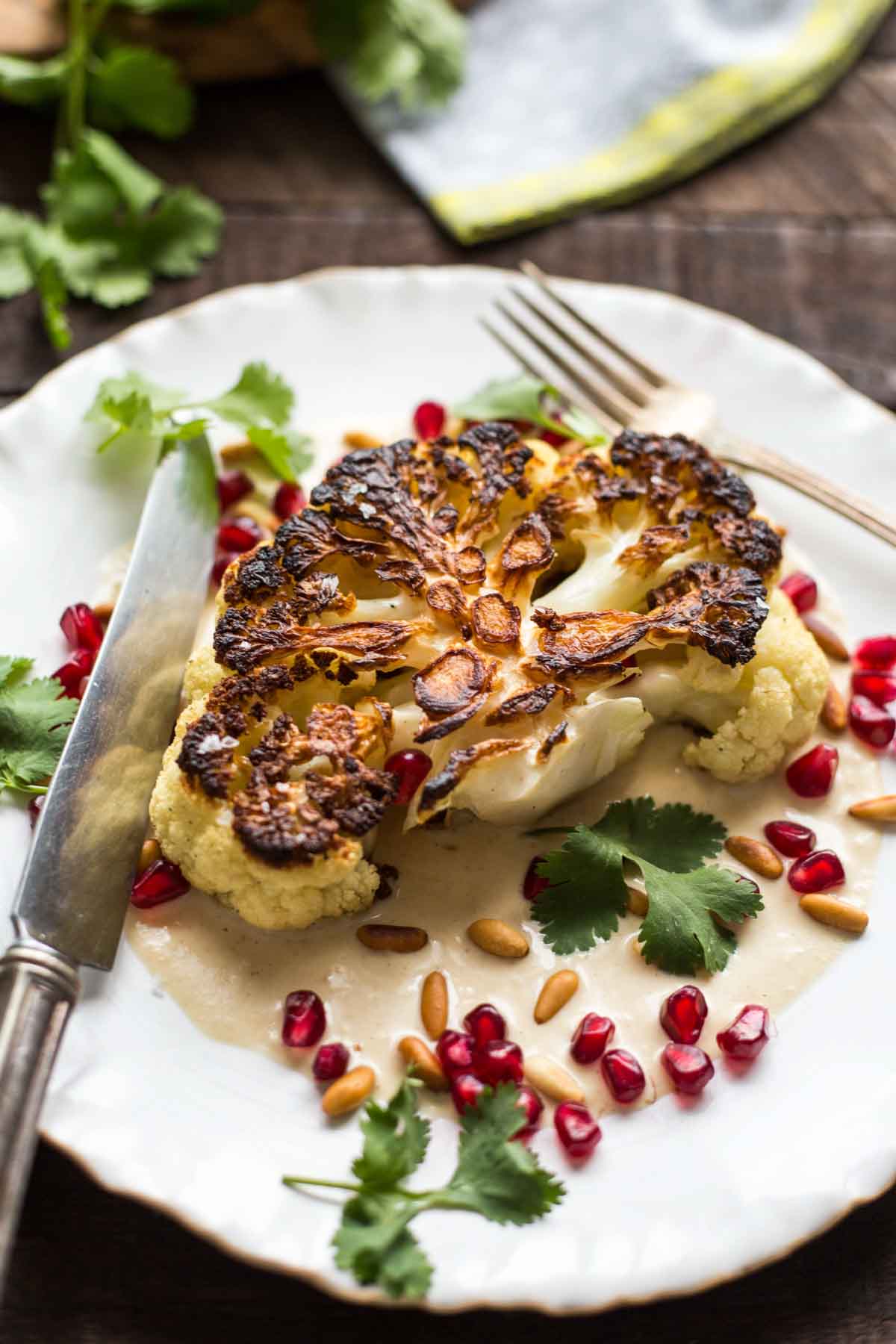 Golden brown cauliflower steak on tahini sauce, garnished with pomegranate arils, cilantro, and pine nuts, photographed with a knife and fork.