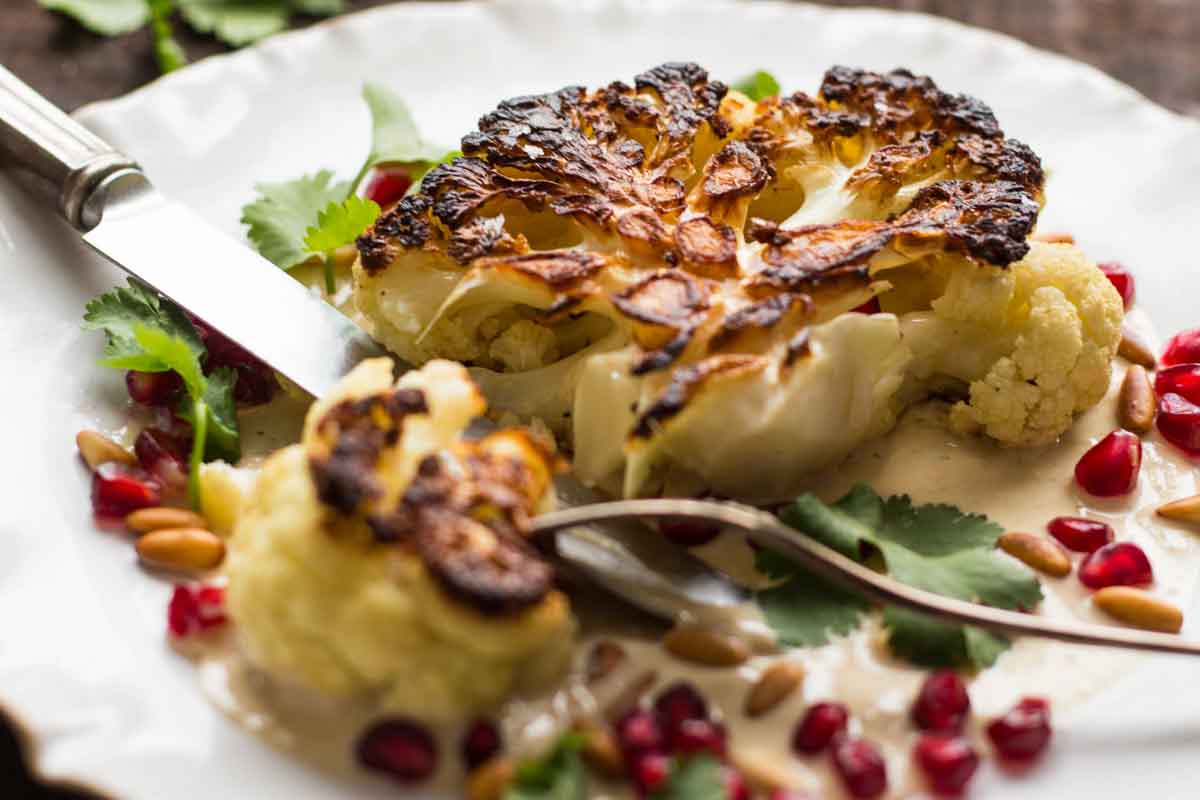 Close-up of a sliced cauliflower steak showing its crisp browned edges and tender center with tahini sauce, pomegranate, and herbs.