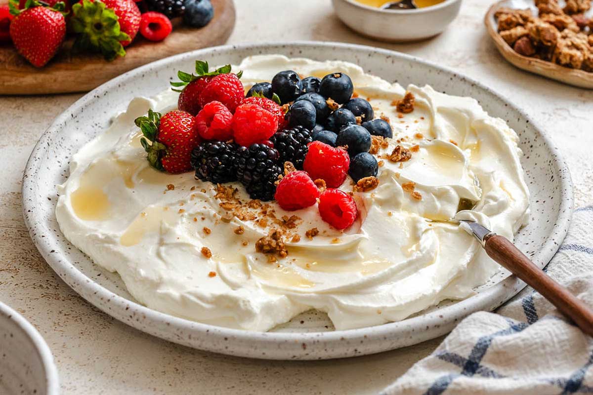 Bowl of thick strained Greek yogurt topped with fresh berries, honey, and granola on a speckled plate.