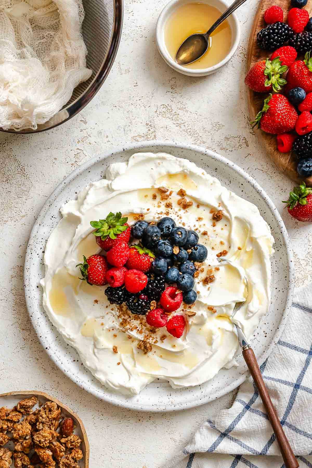 Overhead view of Greek yogurt with mixed berries and honey, surrounded by granola and fresh fruit.
