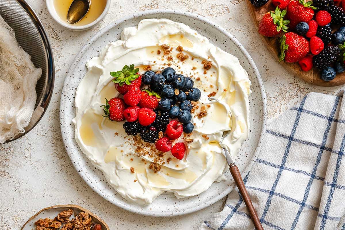 Wide shot of yogurt with berries, honey drizzle, and granola on a textured surface.