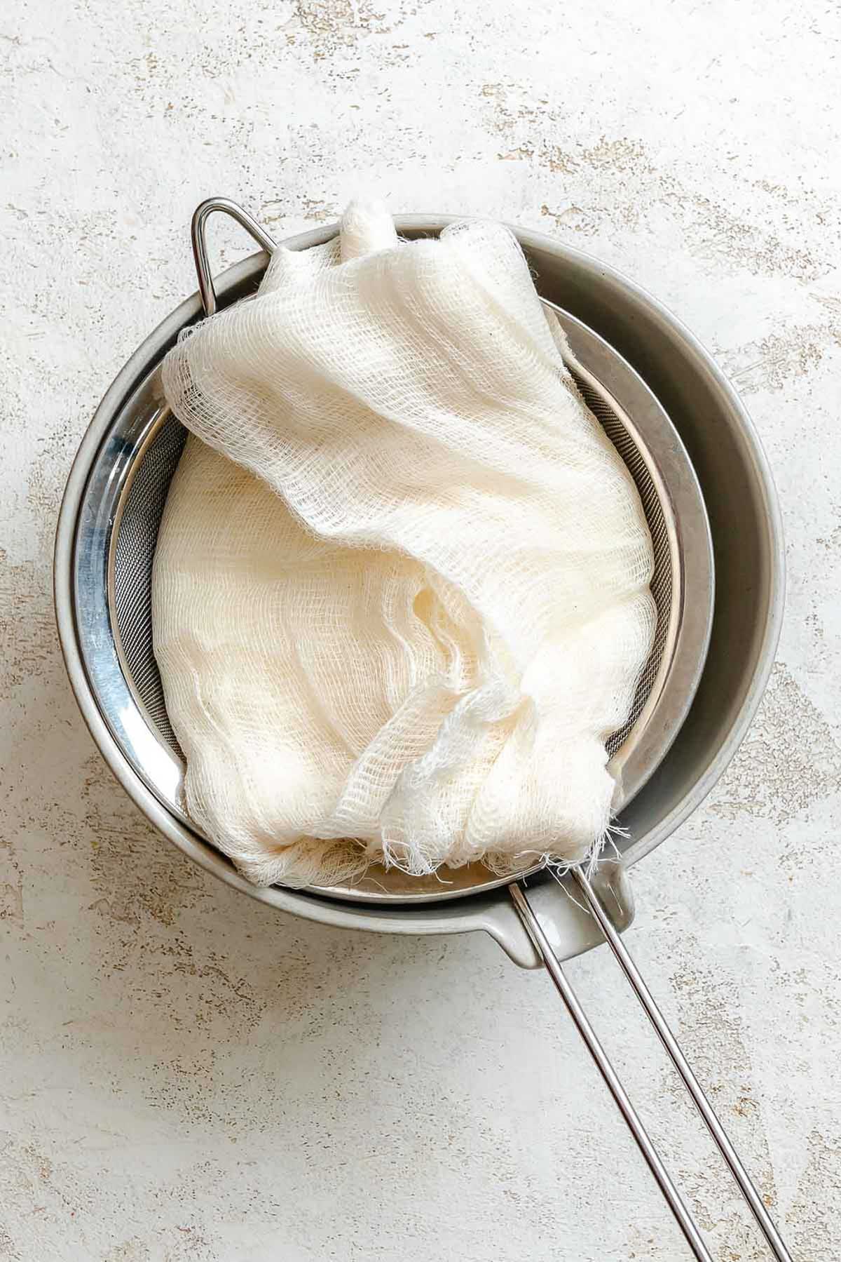 Cheesecloth-wrapped yogurt resting in a metal strainer over a bowl.