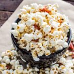 A black bowl filled with truffled popcorn and crispy crumbled prosciutto, surrounded by scattered popcorn on a sheet of parchment paper.