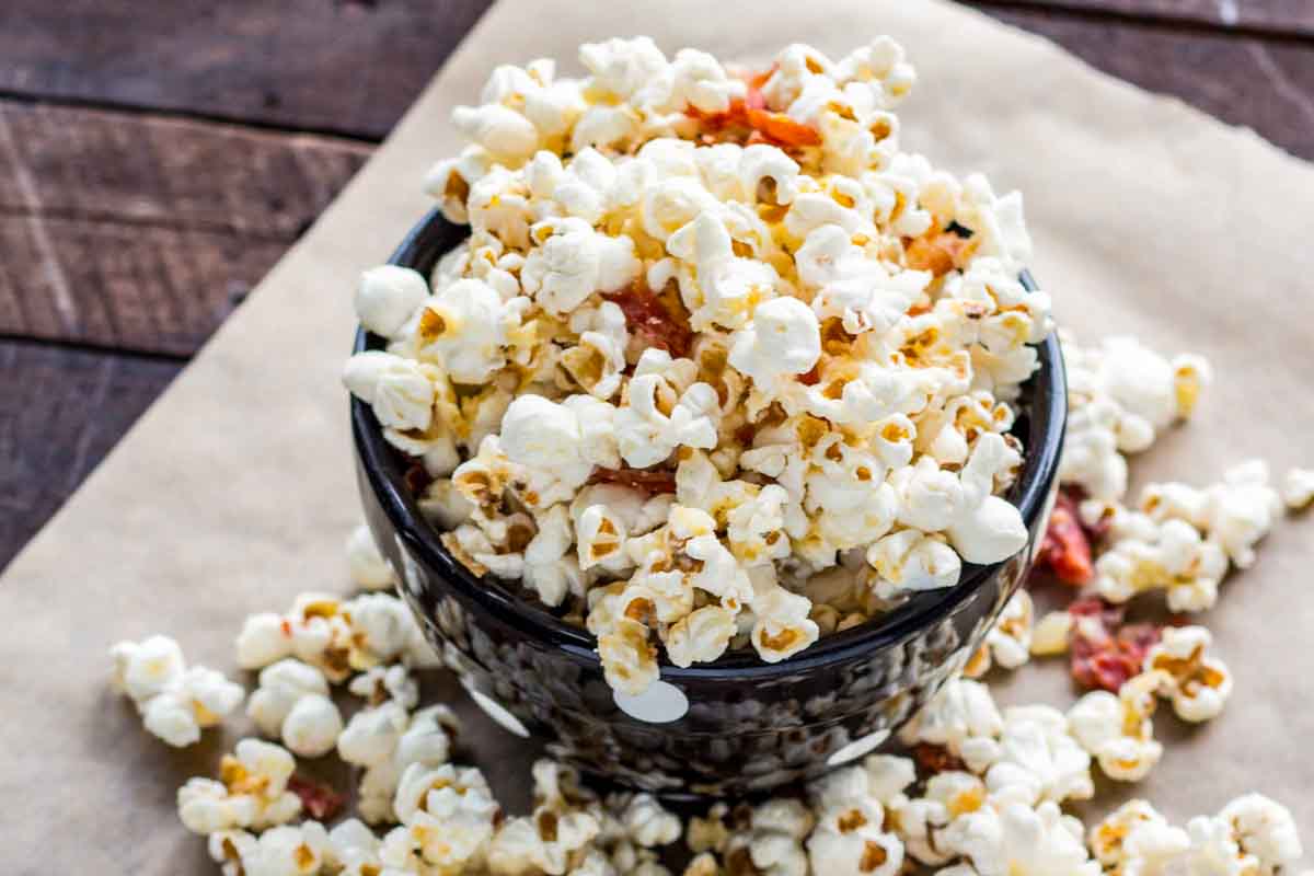 A black bowl filled with truffled popcorn and crispy crumbled prosciutto, surrounded by scattered popcorn on a sheet of parchment paper.