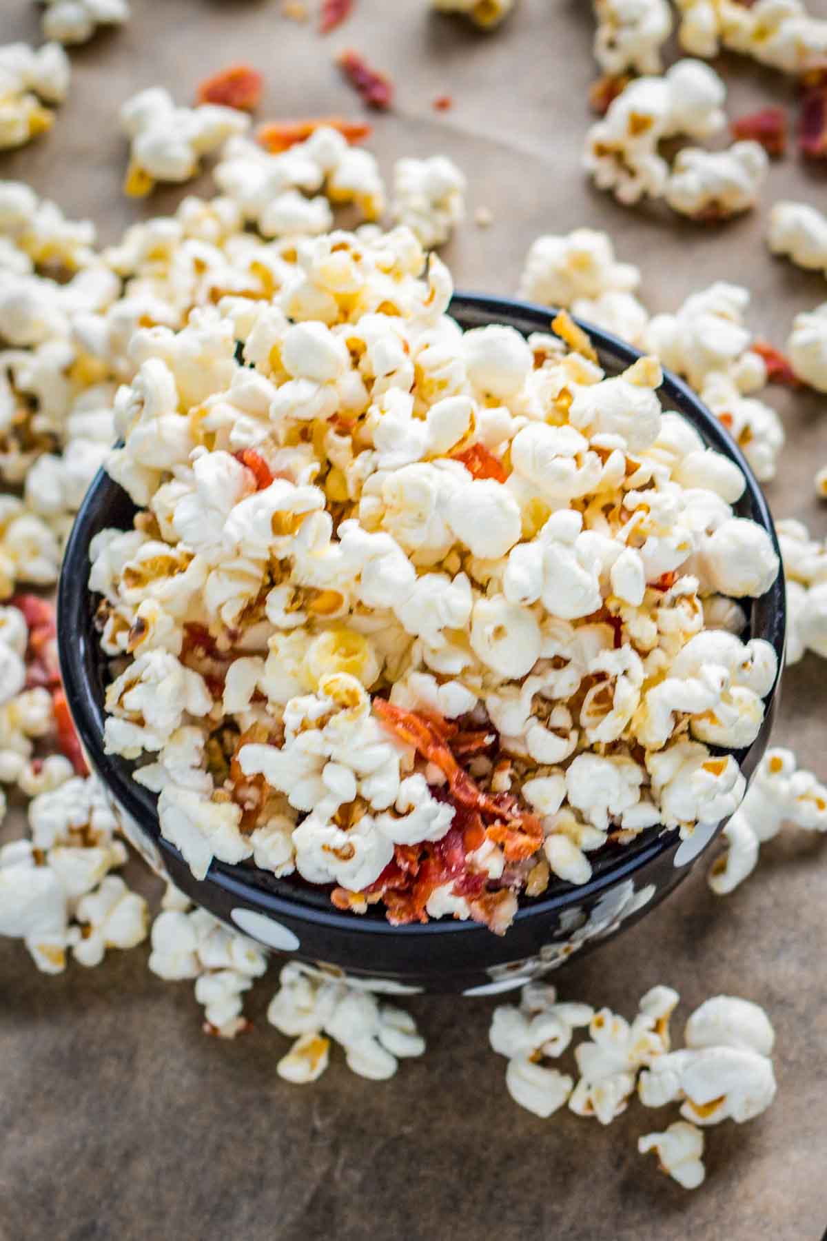 An overhead view of popcorn tossed with crisp prosciutto pieces, piled high in a black bowl with scattered kernels around it.