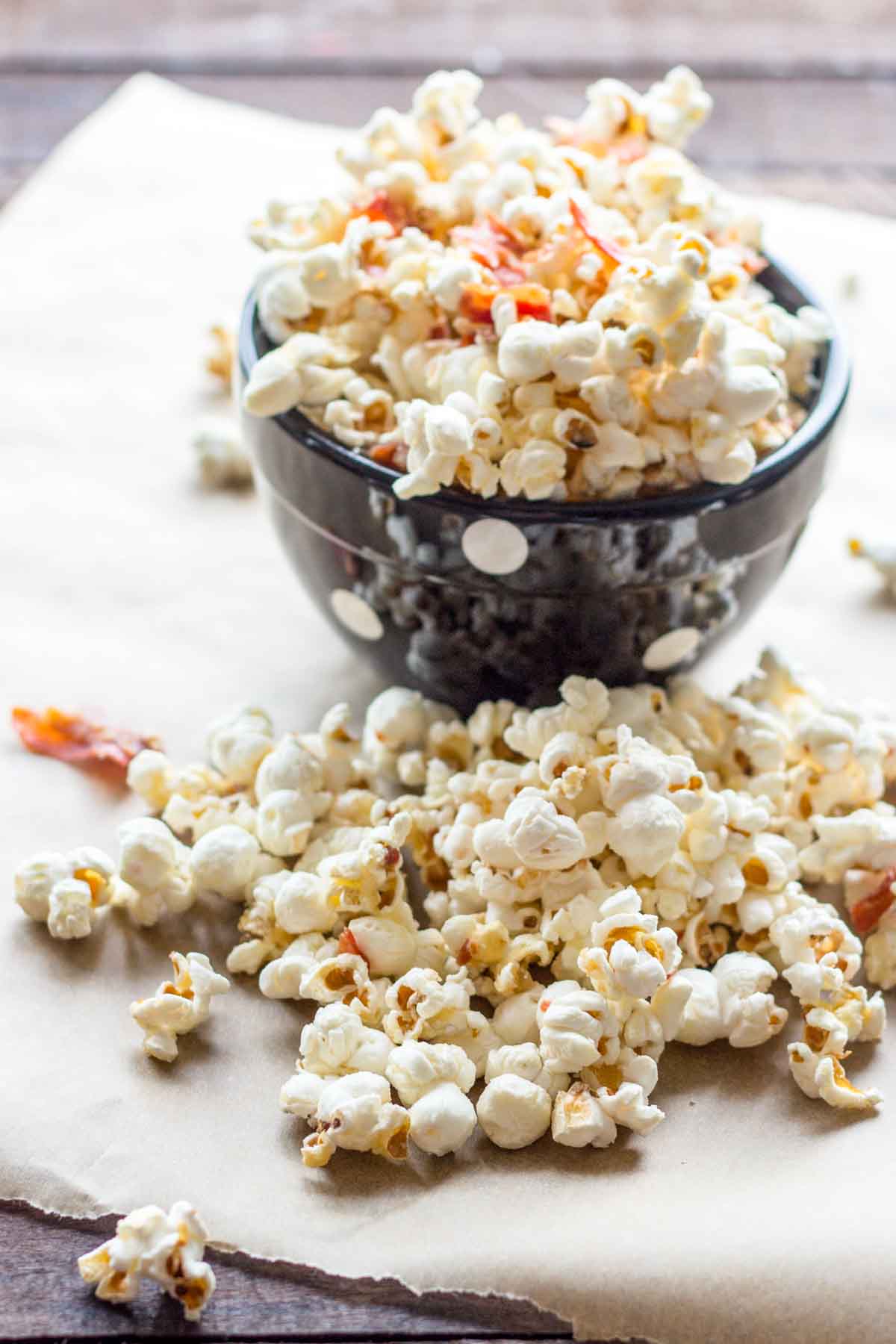 A bowl of popcorn mixed with crunchy prosciutto on parchment paper, with additional kernels and prosciutto pieces scattered in the foreground.