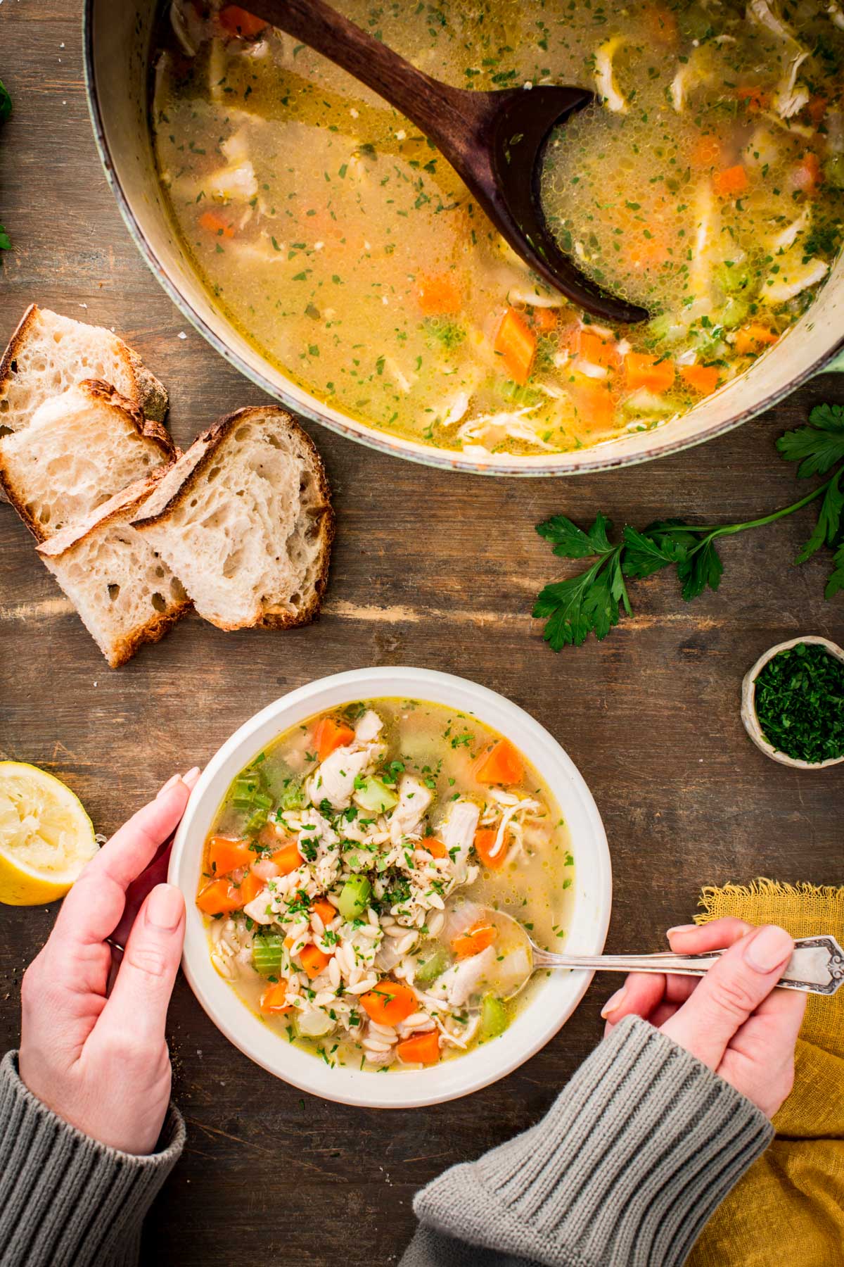 Two hands cradle a bowl of turkey orzo soup next to the pot and sliced bread.