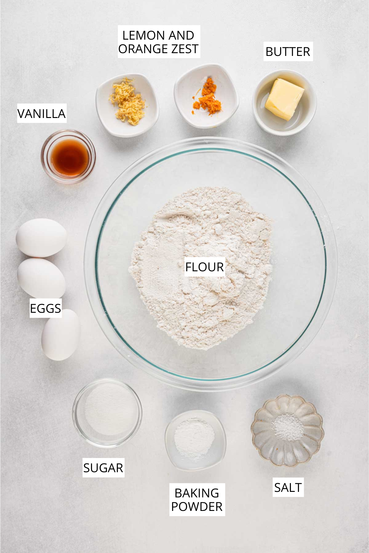A top down view of flour, eggs, sugar, vanilla, butter, baking powder, salt and small bowls of lemon zest and orange zest arranged neatly on a light surface.