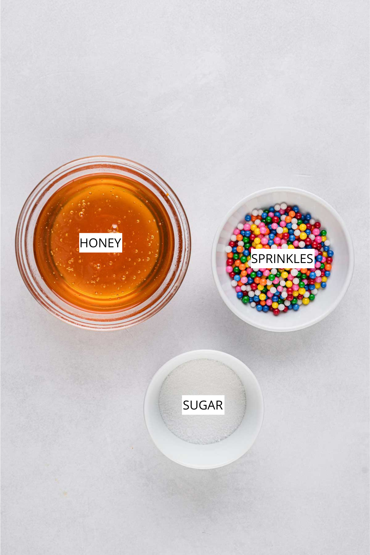A bowl of golden honey next to a dish of colorful round sprinkles and a small bowl of granulated sugar.