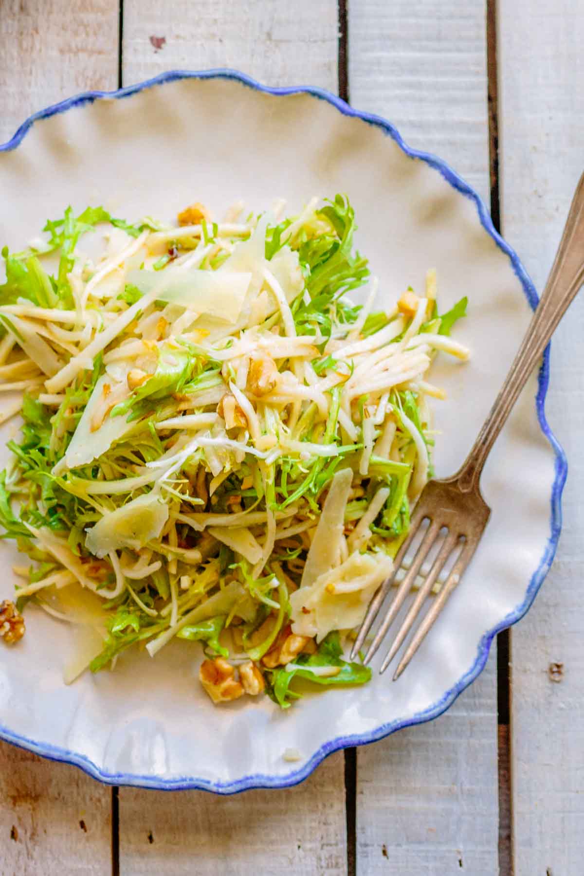 A plate of celery root and pear salad with frisee, walnuts and manchego seen from above on a rustic wooden table.
