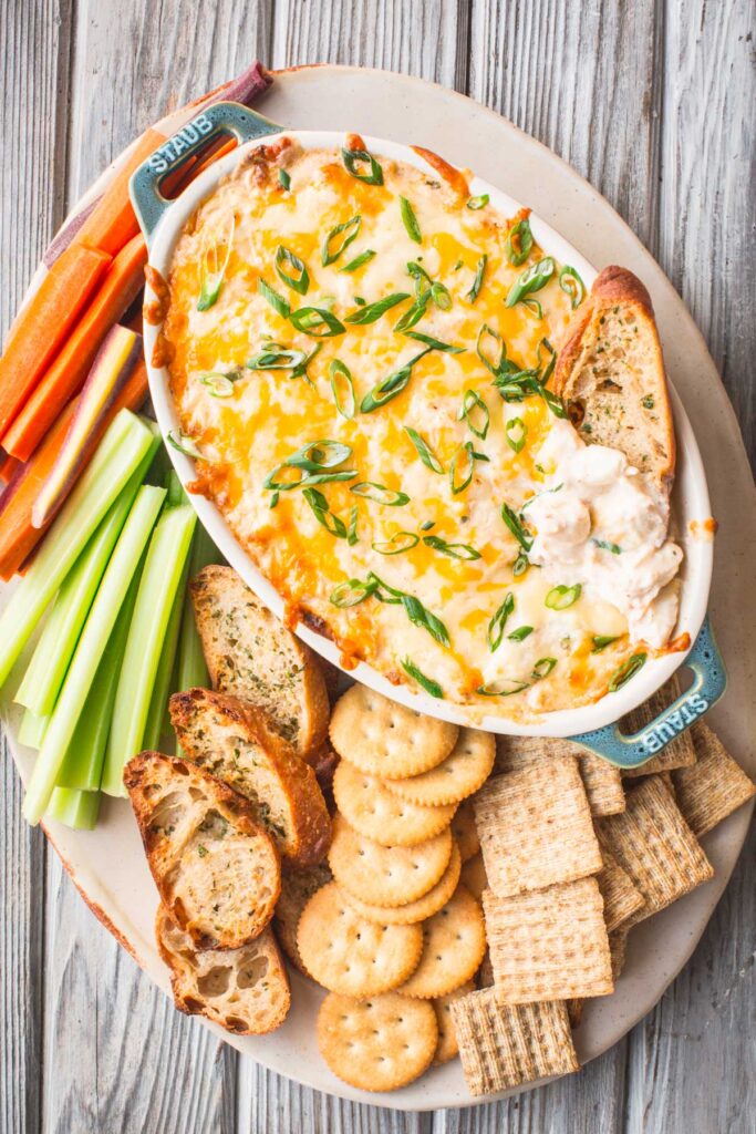 A platter with hot crab dip surrounded by crostini, crackers, celery sticks and carrot sticks.