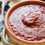 A rustic bowl filled with blended guajillo chili sauce, glossy and deep red, surrounded by dried guajillo chiles and garlic cloves on a wooden surface.